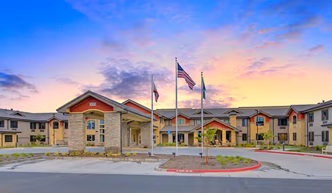 Exterior view of The Pines, a Merrill Gardens community, with flagpoles and a sunset sky.