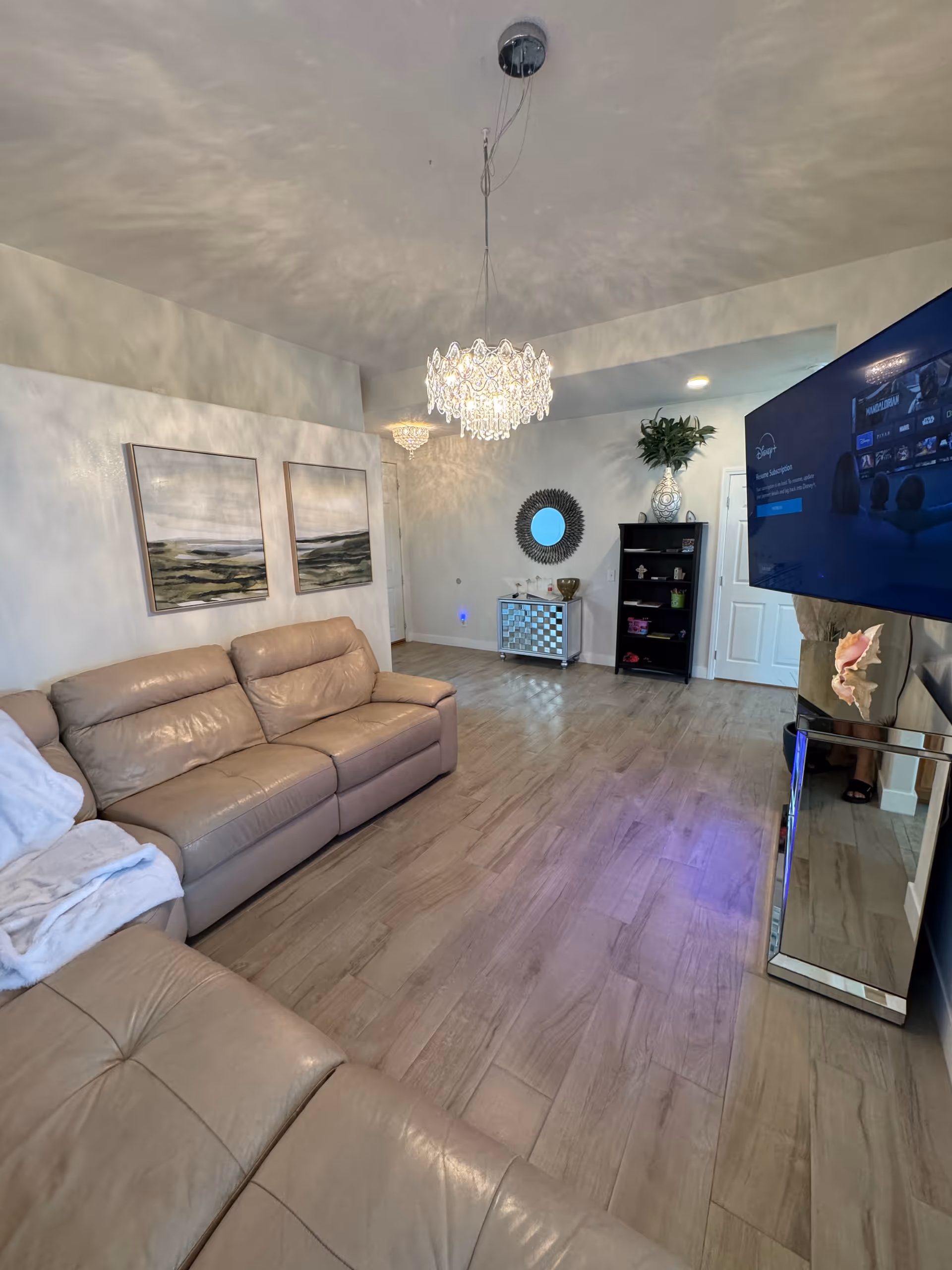 Bright living room with beige sectional, chandelier, TV, and decorative mirrors in a memory care home.