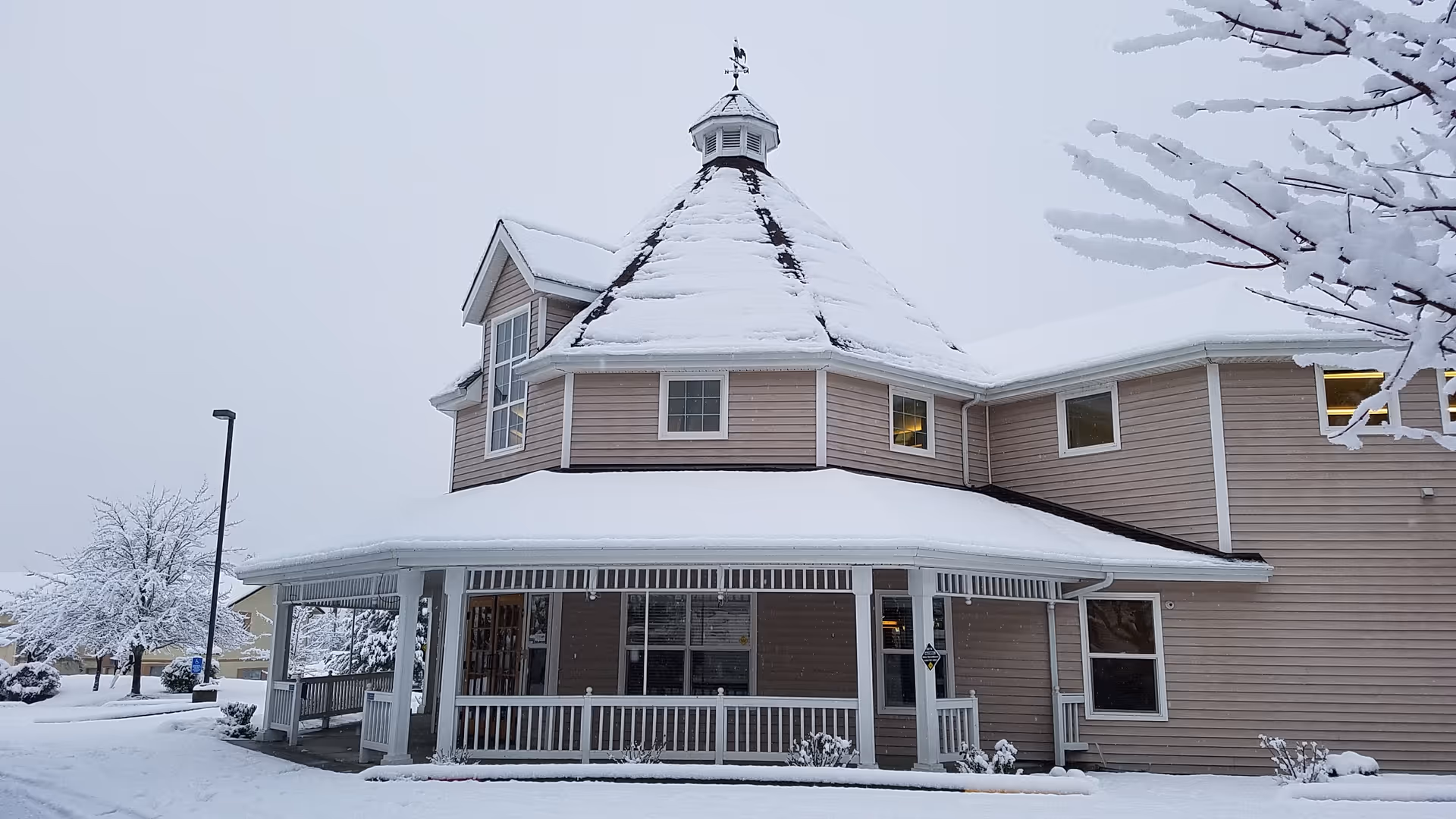 Snow-covered assisted living facility with a wraparound porch and turreted roof.