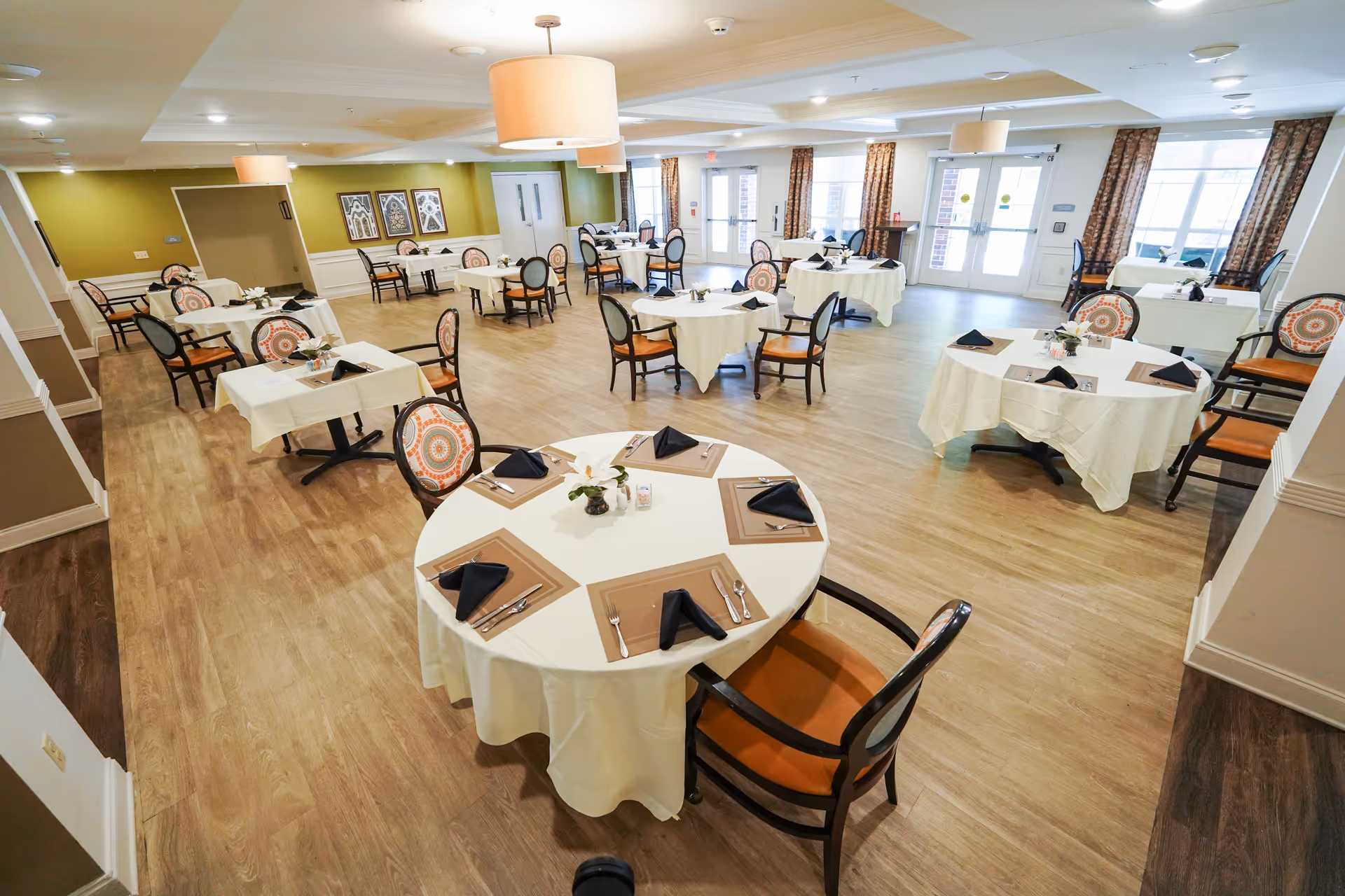 A spacious dining room with multiple round and square tables covered with white tablecloths, each set with placemats, black folded napkins, silverware, and small floral centerpieces. The room has wooden flooring, large windows with patterned curtains, and modern ceiling lights.