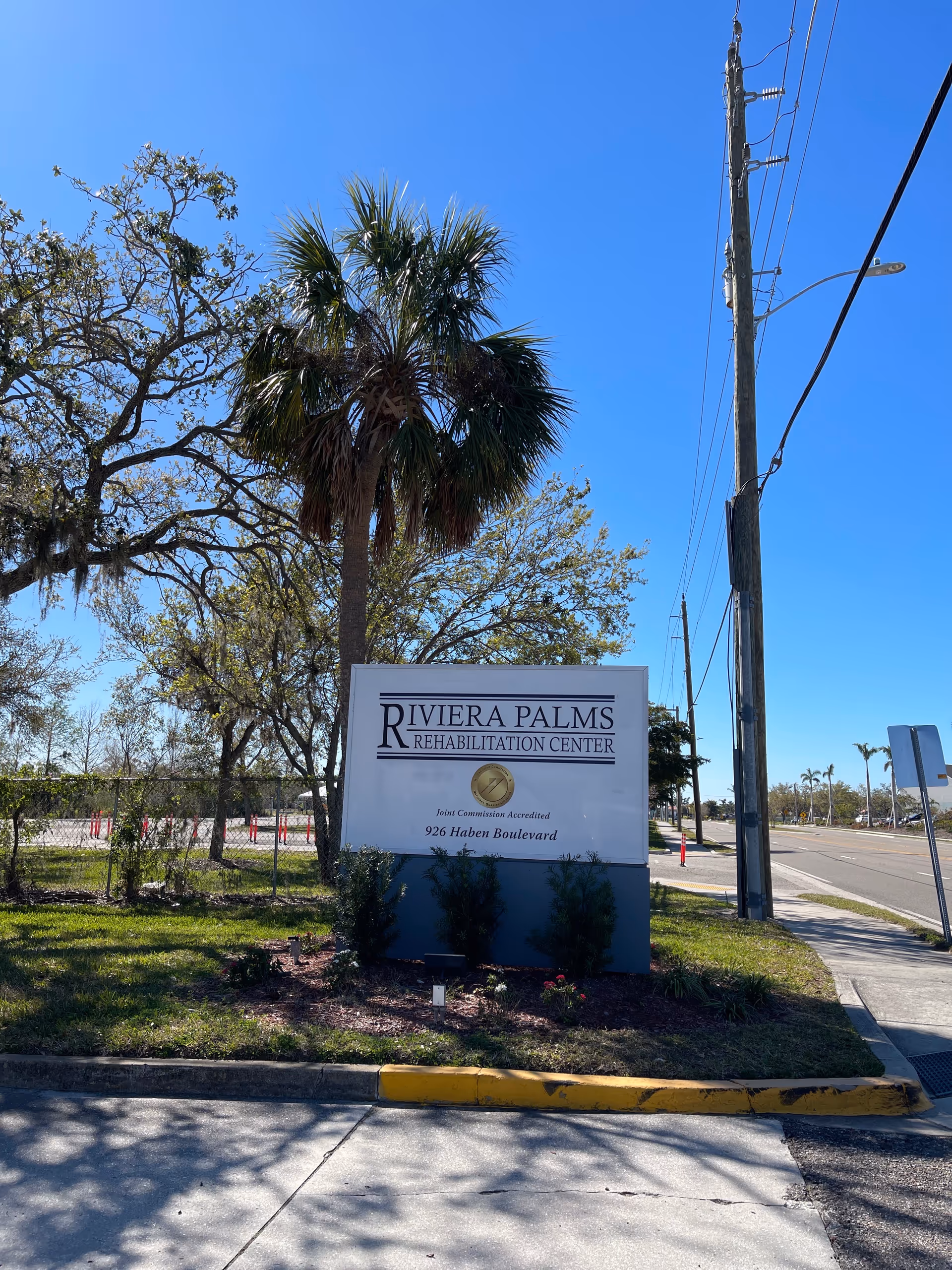 Exterior view of Riviera Palms Rehabilitation Center sign with palm trees and blue sky.