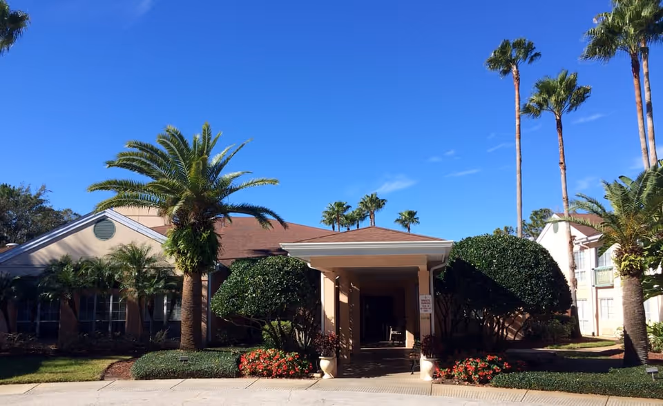 Exterior view of a senior living facility building with a covered entrance, surrounded by palm trees, trimmed bushes, and flower beds under a clear blue sky.