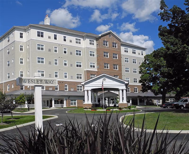 Front exterior of a multi-story senior living building with a porte-cochère, landscaped driveway, and a sign reading "Wesley Way".