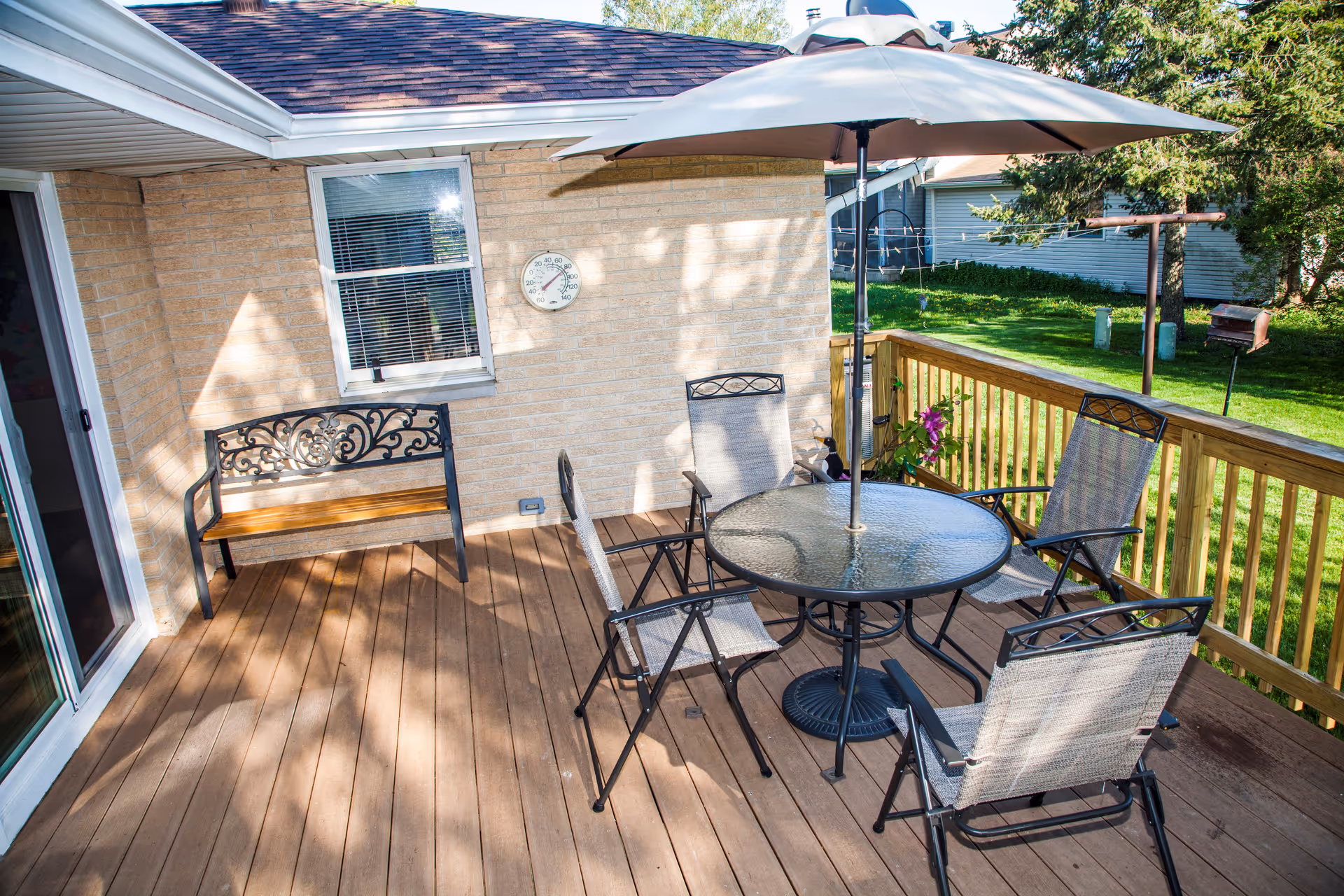 A wooden patio deck with glass table, umbrella, and seating beside a brick house.