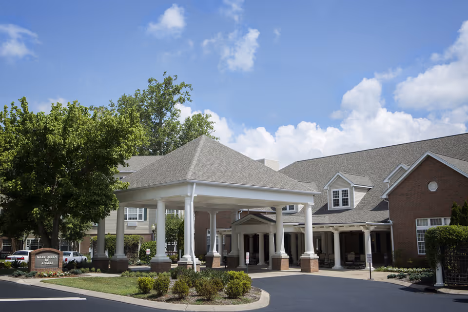 Exterior view of Mary Queen of Angels Assisted living facility showing a covered entrance with white columns, a brick building, green trees, and a clear blue sky with some clouds.