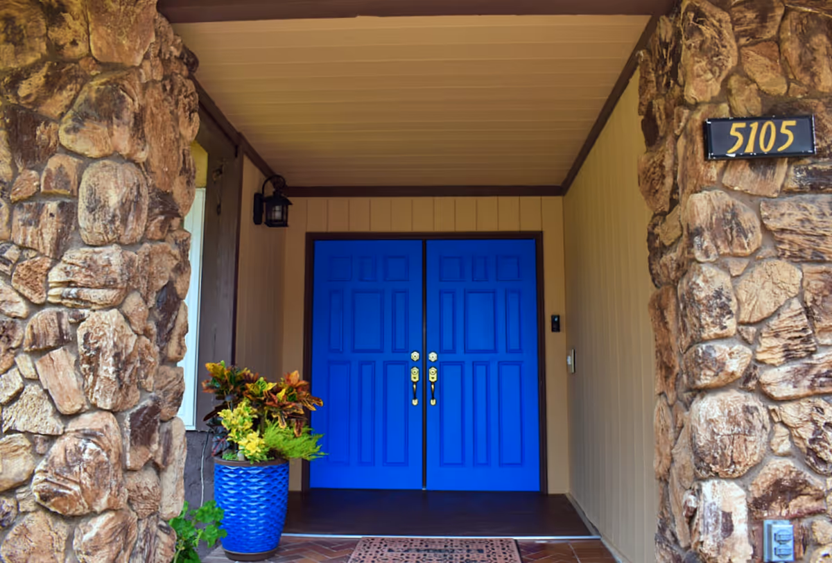 Entrance to a building with a pair of bright blue double doors, flanked by stone walls. There is a blue planter with green and yellow foliage on the left side of the entrance, a small outdoor light fixture above the door, and the number 5105 displayed on the right stone wall.
