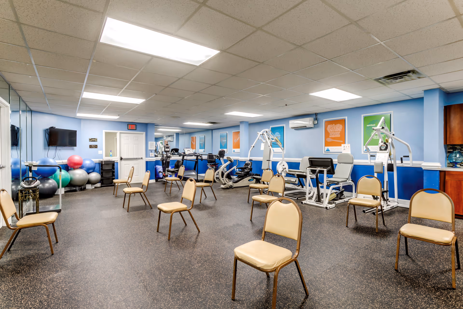 A fitness room with exercise equipment including stationary bikes, weight machines, and a rack of colorful exercise balls. Several beige chairs are spaced out across the room. The walls are painted blue with motivational posters, and there is a wall-mounted TV and mirrors on one side.