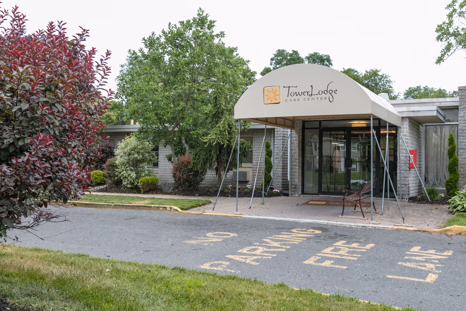 Tower Lodge Care Center entrance with a beige canopy over glass doors and surrounding greenery.