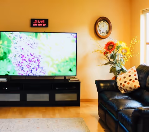 A cozy living room with a black leather couch adorned with a white pillow with brown polka dots. A large flat-screen TV on a black TV stand displays a close-up image of purple flowers. Above the TV, a digital clock shows the time and date. A wall clock and a vase with colorful artificial flowers are placed on the wall and floor respectively, next to the couch. Light wooden flooring and a window letting in natural light complete the scene.