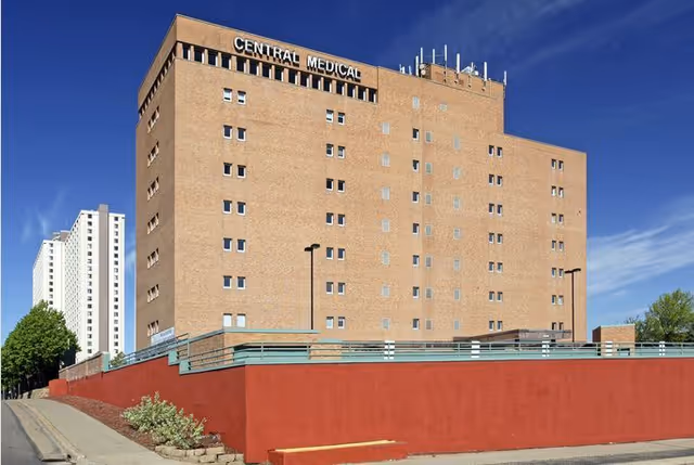 Tall tan brick medical building with a red retaining wall, metal railing, and clear blue sky.