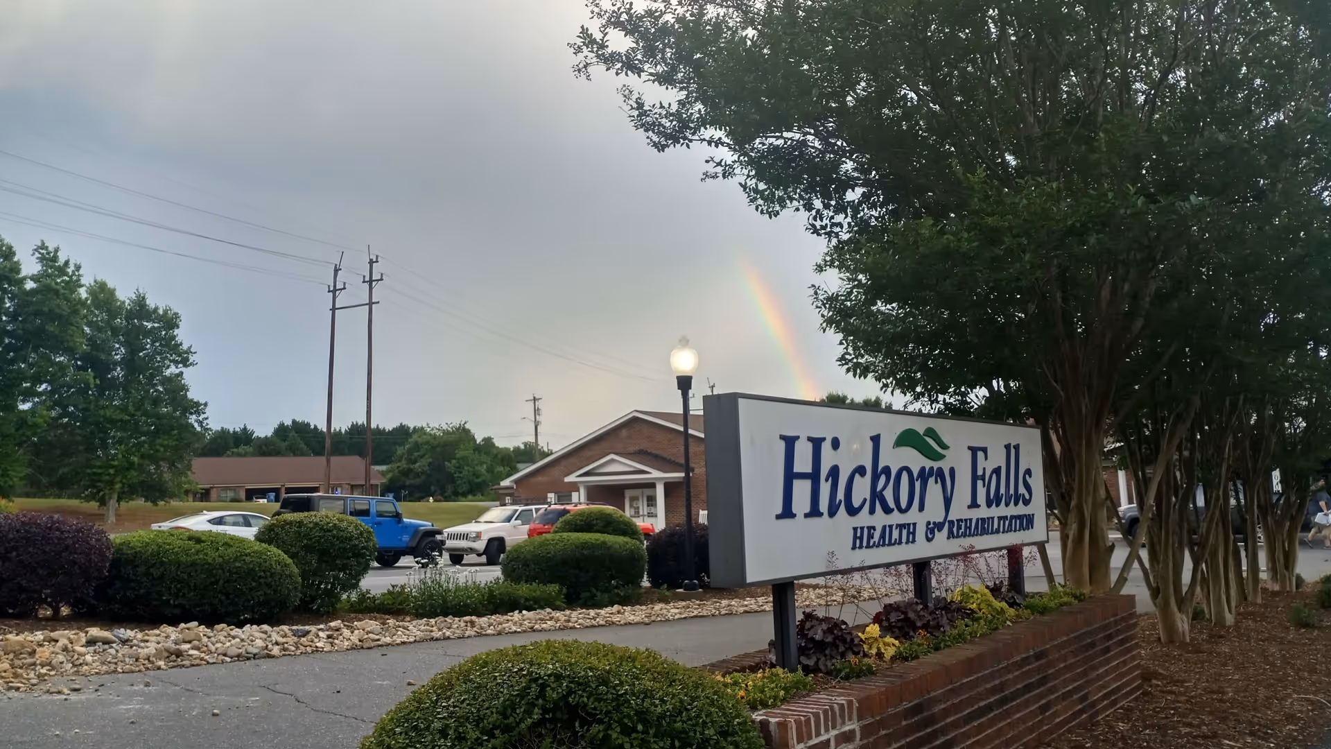 Exterior view of Hickory Falls Health and Rehabilitation sign with parking and rainbow.