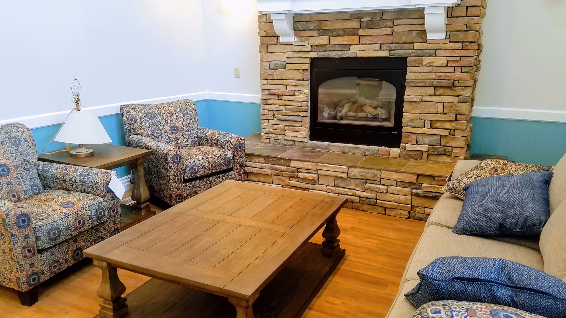 A cozy living room area featuring a stone fireplace with a wooden mantel, two patterned armchairs, a wooden coffee table, a side table with a lamp, and a beige sofa with blue and patterned cushions.