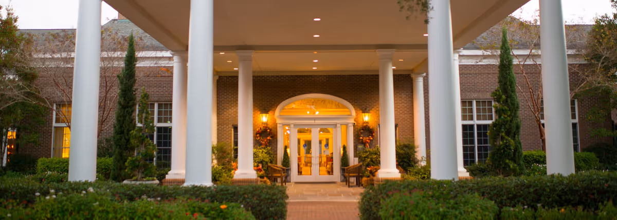 Entrance of a brick building with large white columns and a covered walkway. The doorway is decorated with wreaths and flanked by plants and benches. The building is surrounded by greenery and trees.