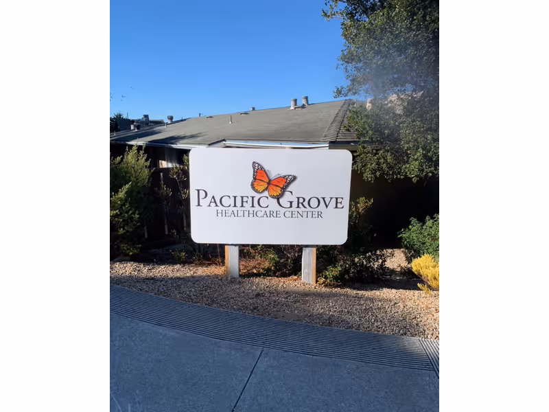 Outdoor view of a sign for Pacific Grove Healthcare Center with a butterfly logo, set in a landscaped area with bushes and gravel, and a building with a sloped roof in the background under a clear blue sky.