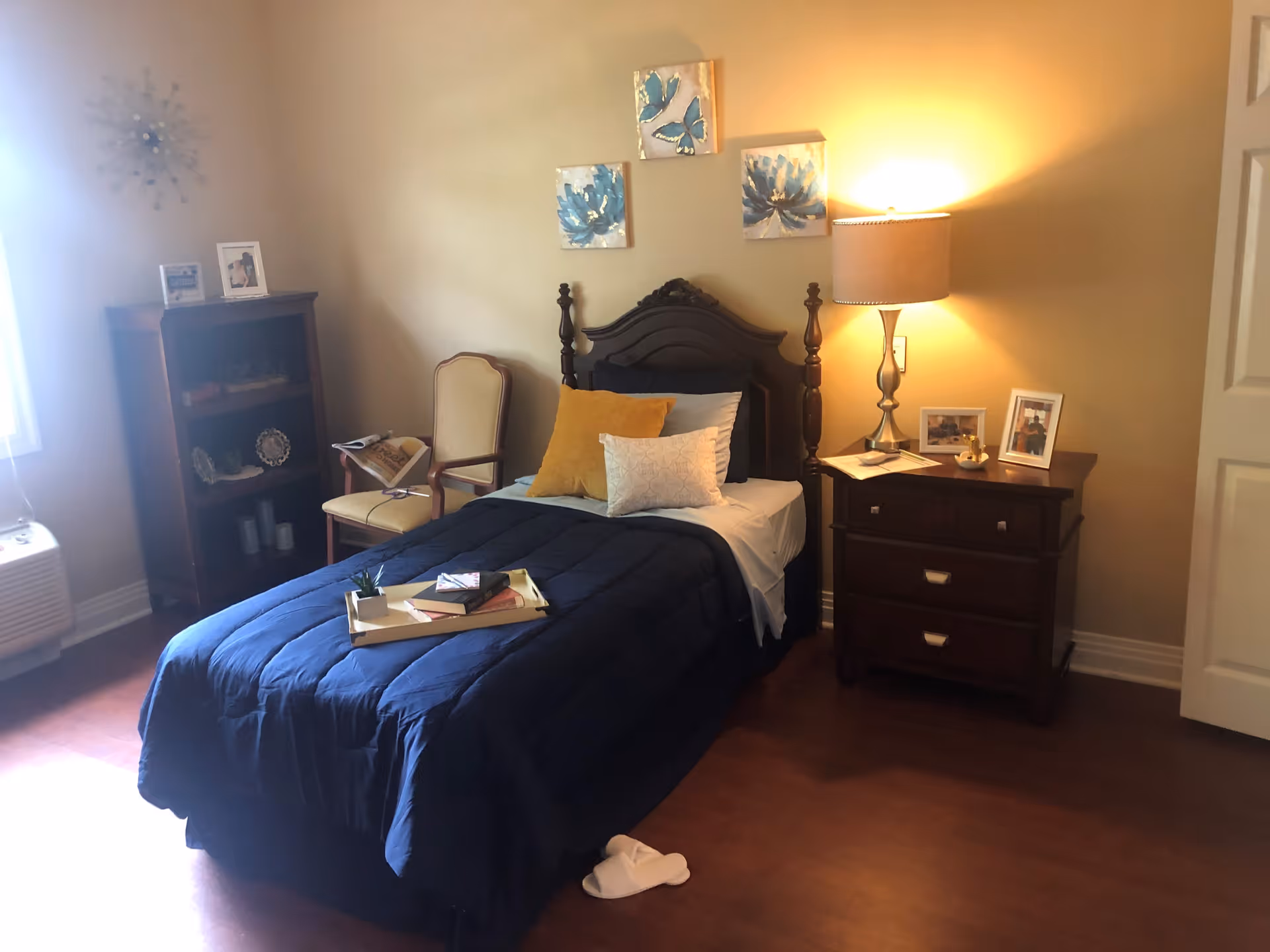 A cozy bedroom with a single bed covered in a dark blue comforter and several pillows. A wooden nightstand with a lamp, framed photos, and a small decorative item is beside the bed. There is a wooden chair with a cushion and a small bookshelf with decorative items and framed photos. The room has warm lighting and wooden flooring.