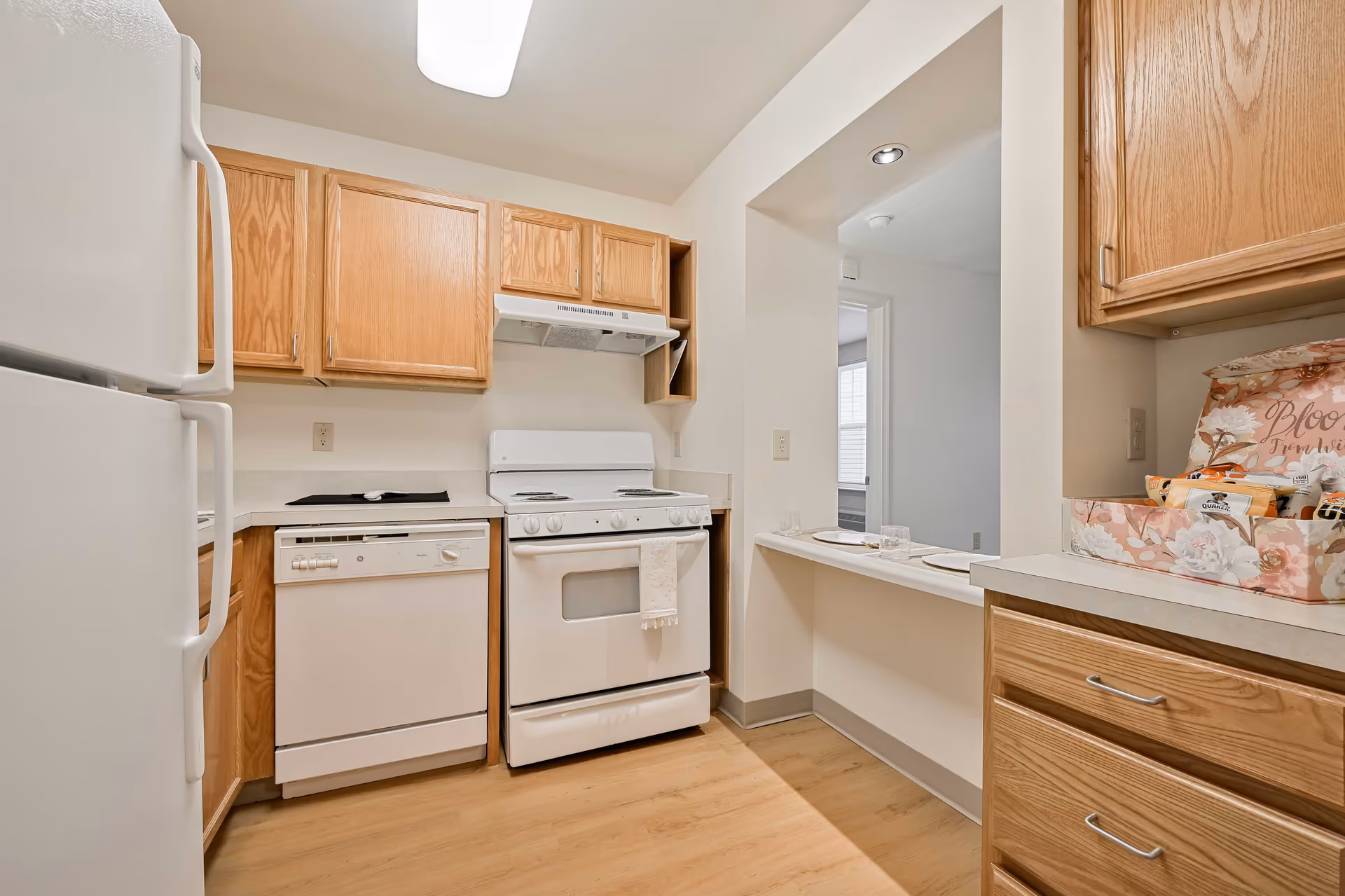 Compact kitchen with white appliances, wooden cabinets, and a pass-through to another room.