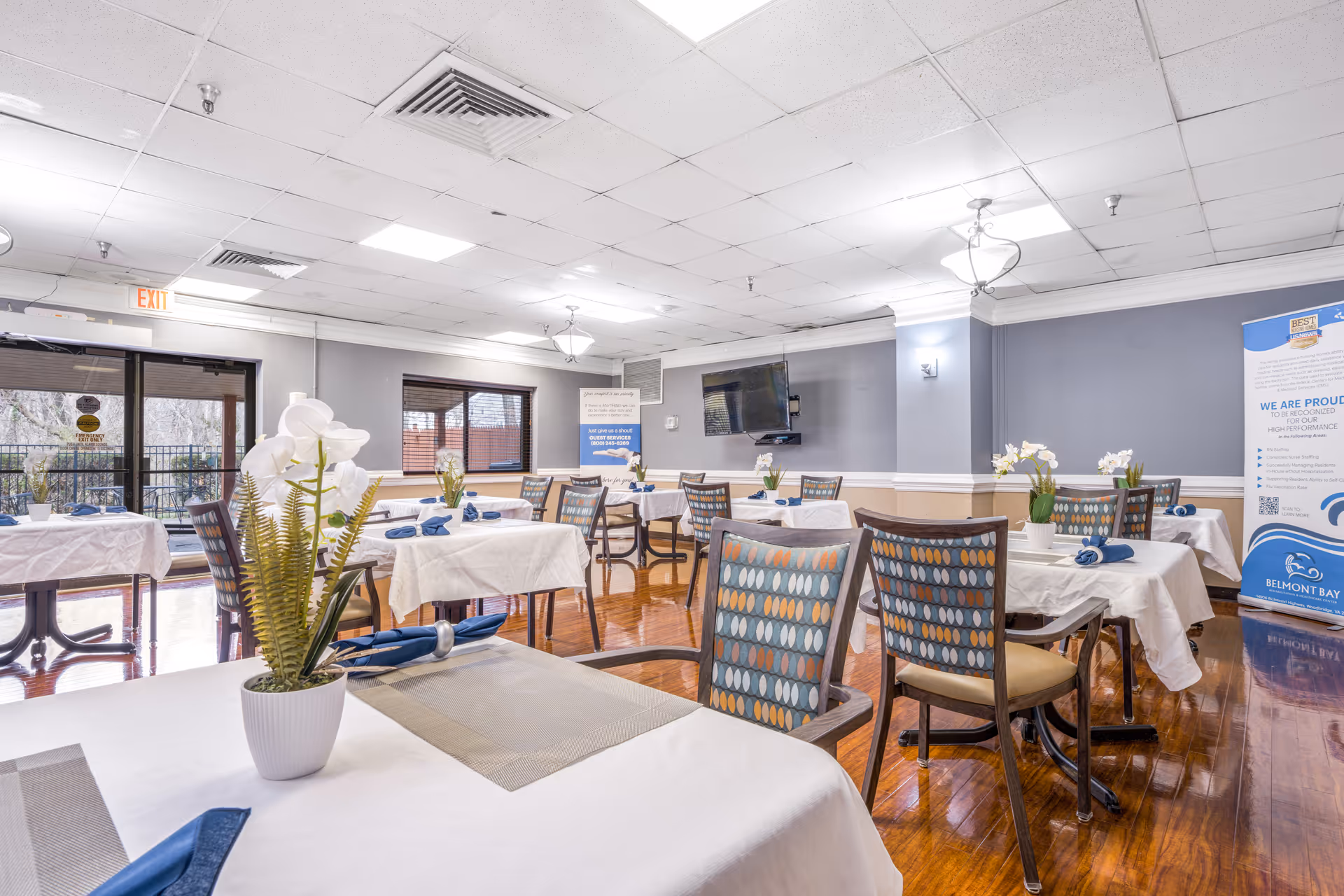 Bright senior living dining room with white tablecloths, blue napkins, and flower centerpieces.
