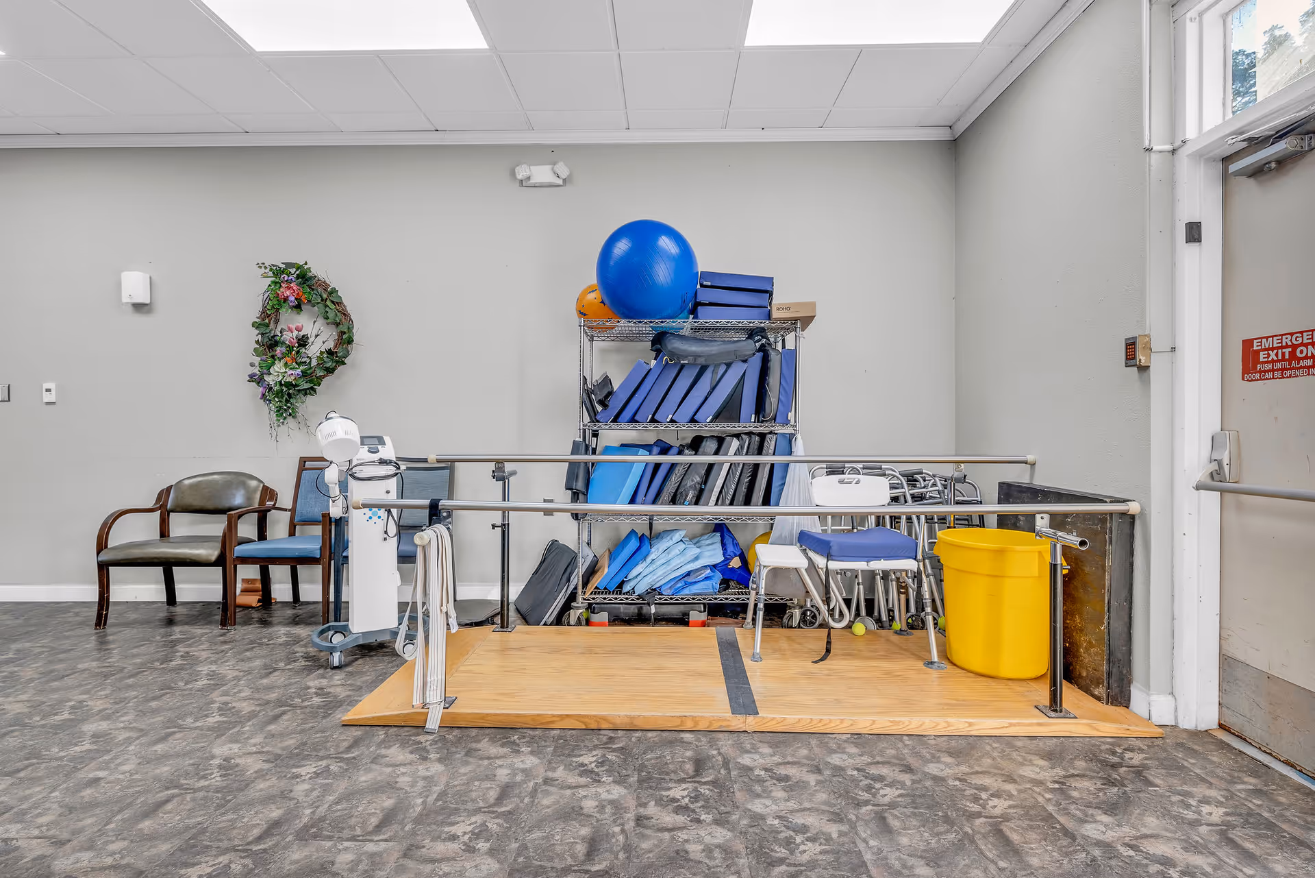 Physical therapy area in a rehabilitation center with parallel bars on a wooden platform, a yellow bucket, chairs, a rack filled with blue exercise mats and therapy equipment, and an emergency exit door on the right.