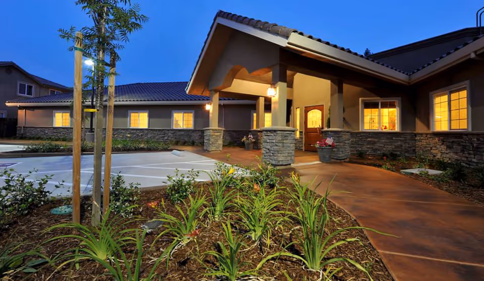 Exterior view of The Meadows at Country Place facility at dusk, showing a well-lit entrance with stone pillars, a covered walkway, and landscaped garden beds with plants in the foreground.