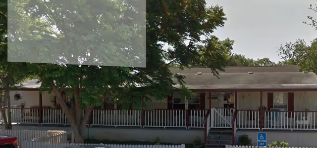 Single-story care home exterior with a long covered porch, railing, steps, and surrounding trees.