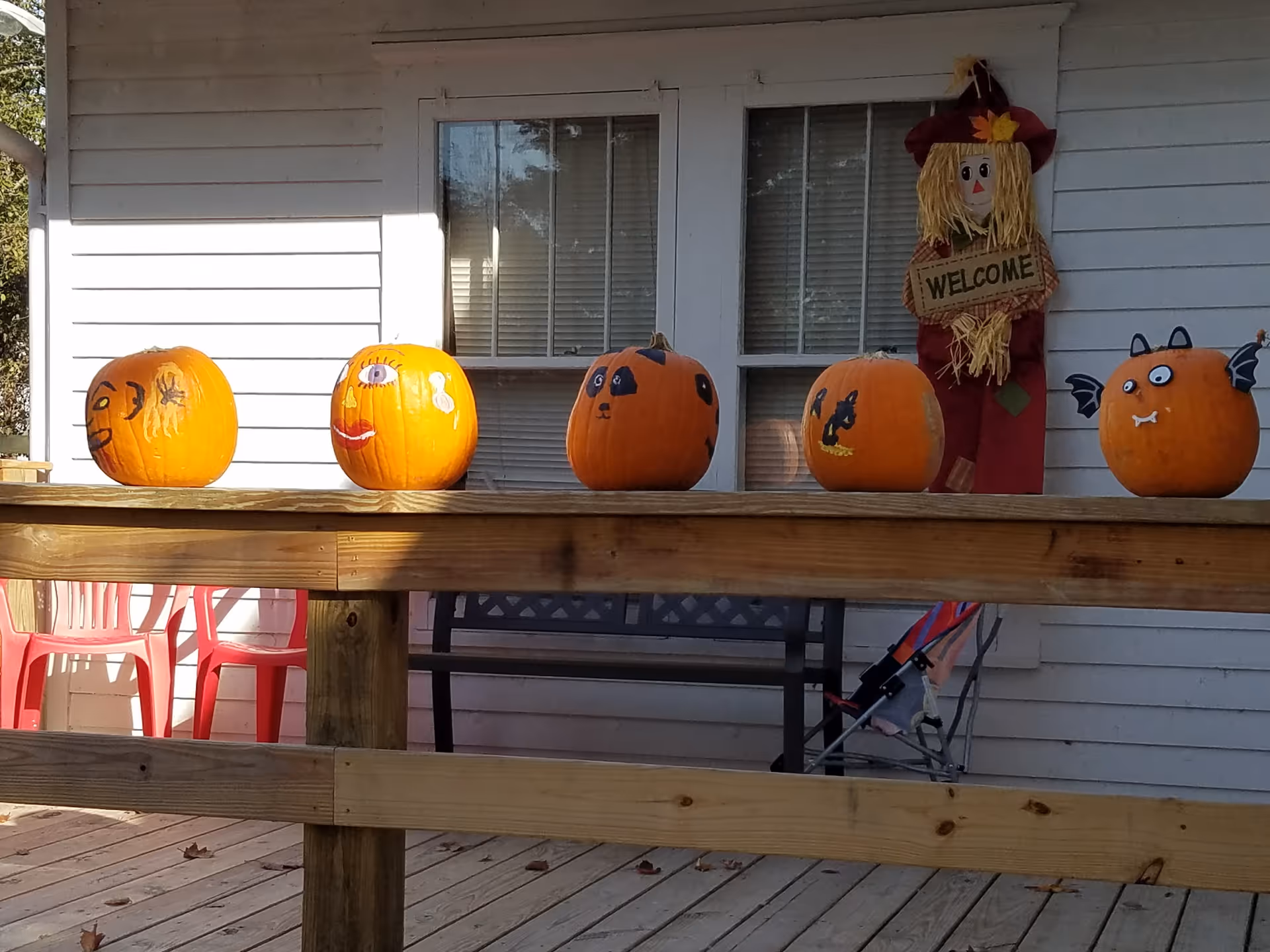 Pumpkins with painted faces sit along a wooden porch railing outside a white house.