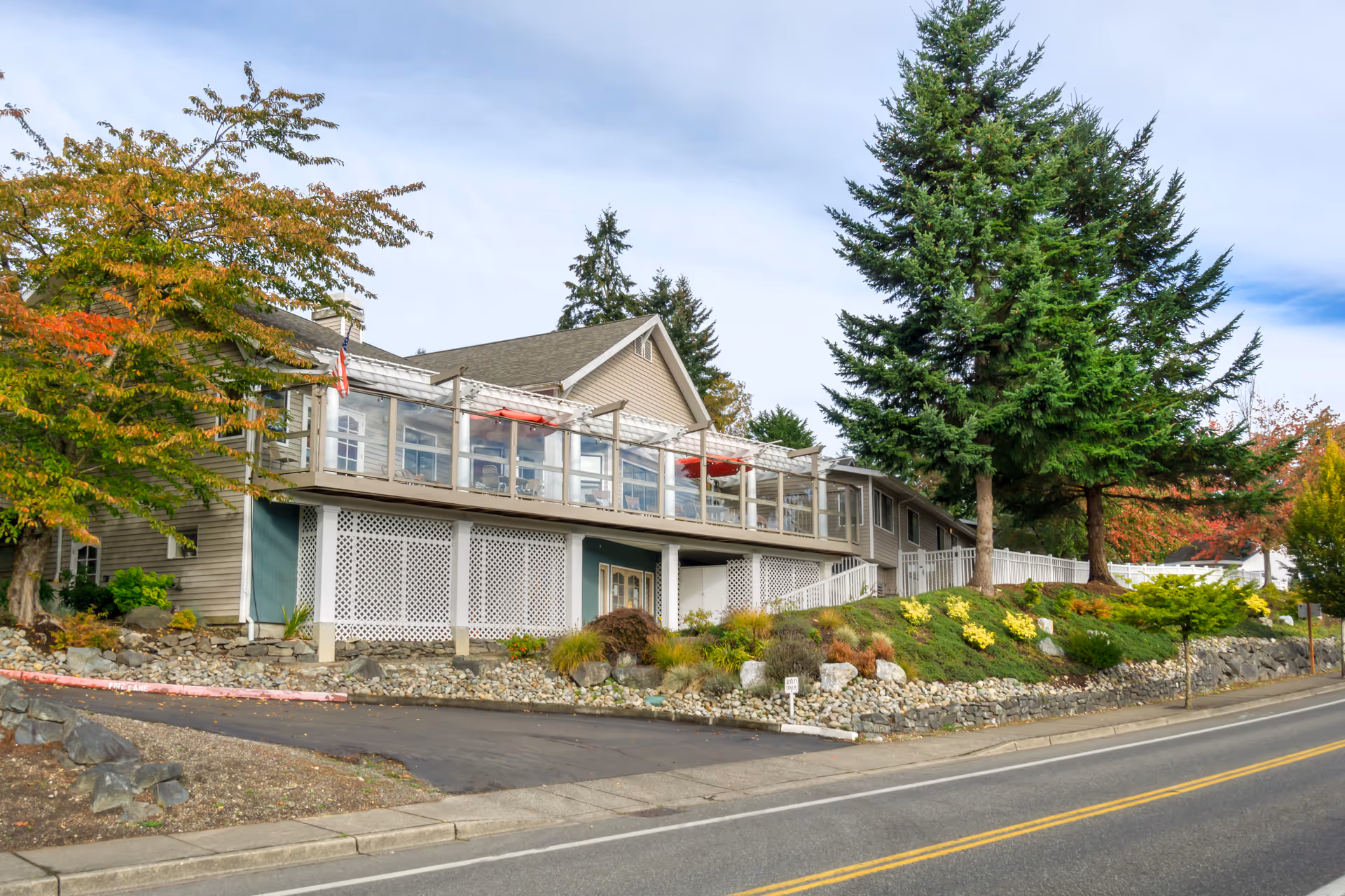 Two-story senior living facility with glass-railed deck, landscaped front, and surrounding trees.