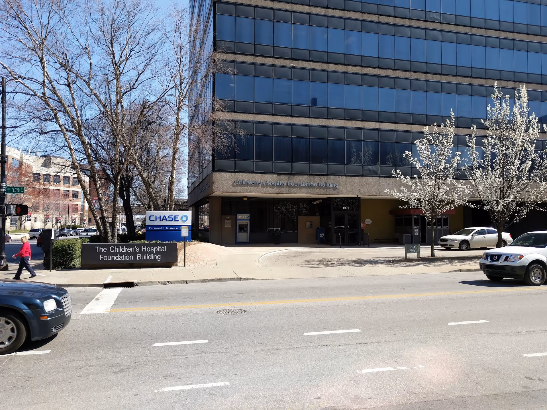 Tall glass-front building on a city street with blooming trees and cars