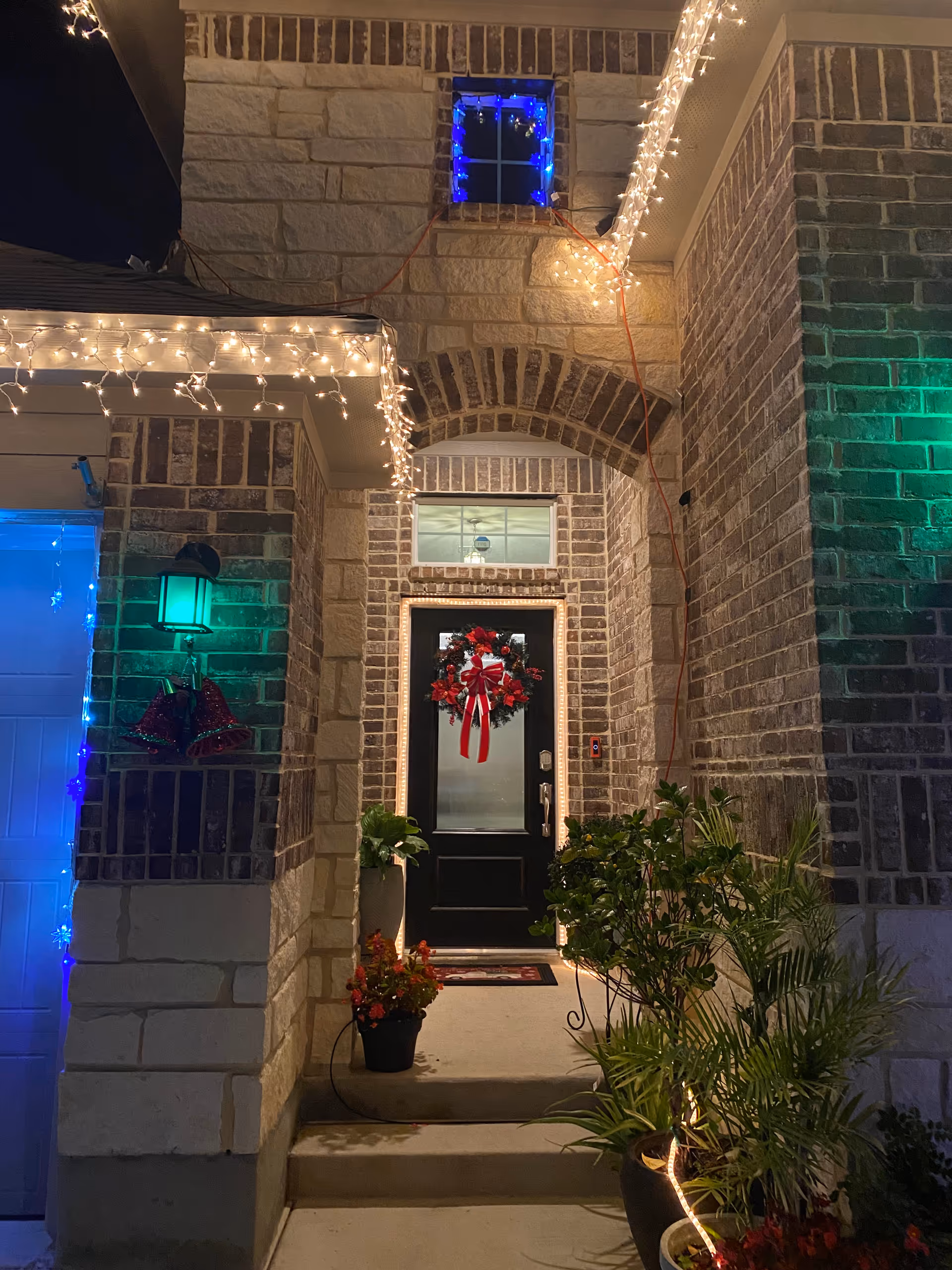 Night exterior of a brick building with festive lights, wreath, and potted plants at the entrance.