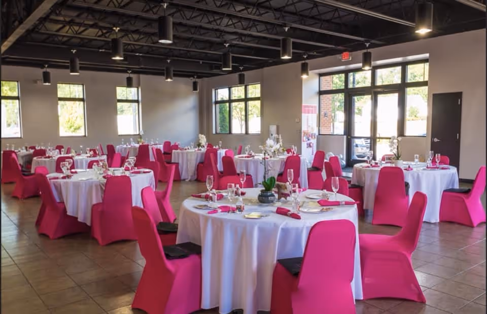 Inside a bright dining hall, round tables with white linens and pink chair covers.