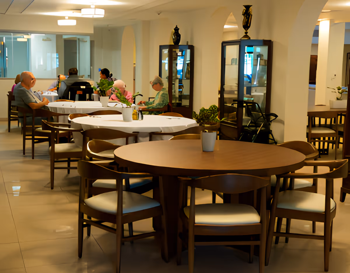 A communal dining room with round wooden tables, chairs, and several elderly residents seated and conversing.