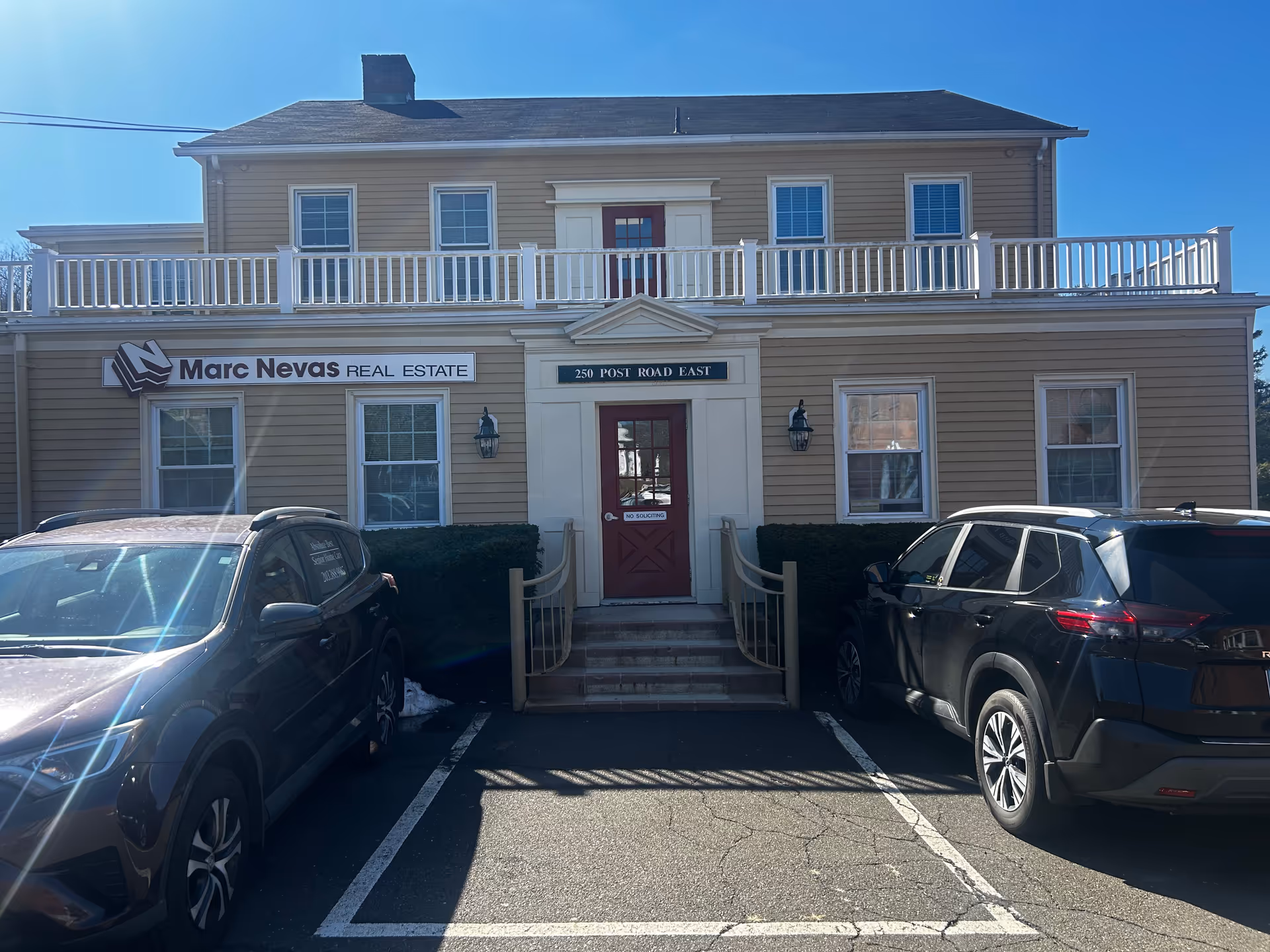 Two-story beige building with white trim, a red front door, and cars parked in front.