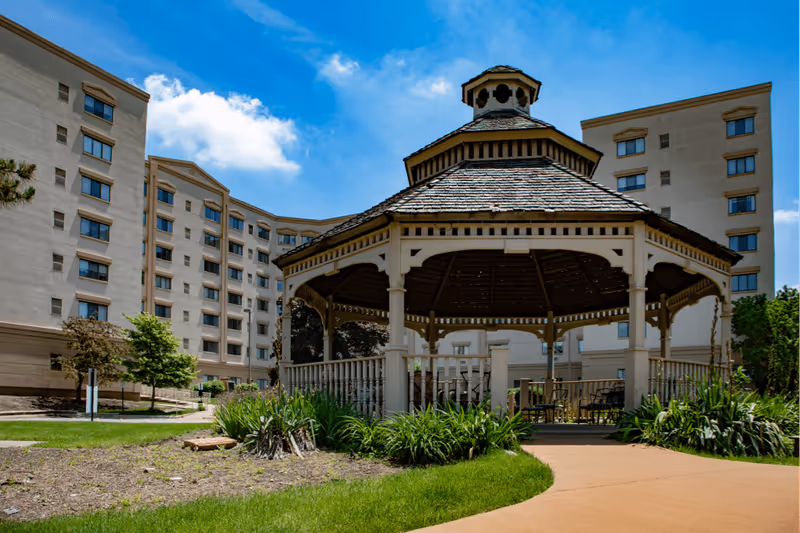 A wooden gazebo in a landscaped courtyard in front of a multi-story senior living building under a blue sky.