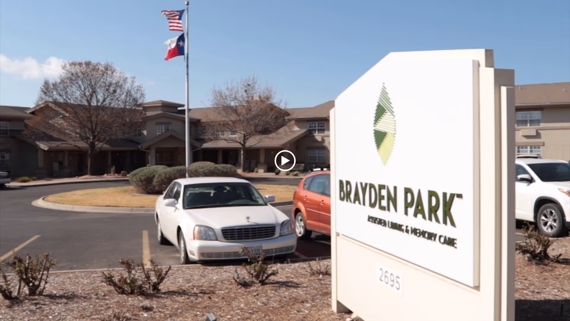 Exterior view of Brayden Park Assisted Living & Memory Care facility with a large sign in the foreground displaying the facility name and address number 2695. Several cars are parked in front of the building, and two flagpoles with the American and Texas flags are visible. The building is surrounded by some landscaping and trees under a clear blue sky.