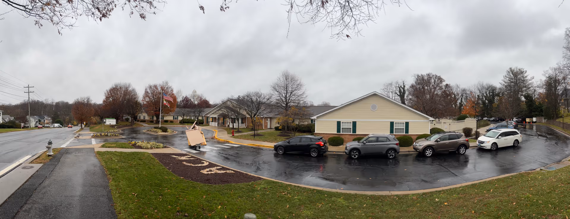 Panoramic exterior view of a memory care community building with a flagpole, wet driveway and several parked cars on a cloudy day.