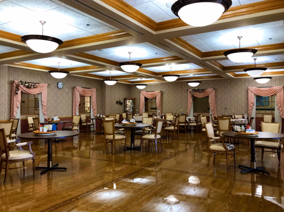 A spacious dining room with multiple round tables and chairs arranged neatly. The room features wooden floors, patterned wallpaper, and windows adorned with pink drapes and floral decorations. Ceiling lights with wooden beams provide bright illumination.