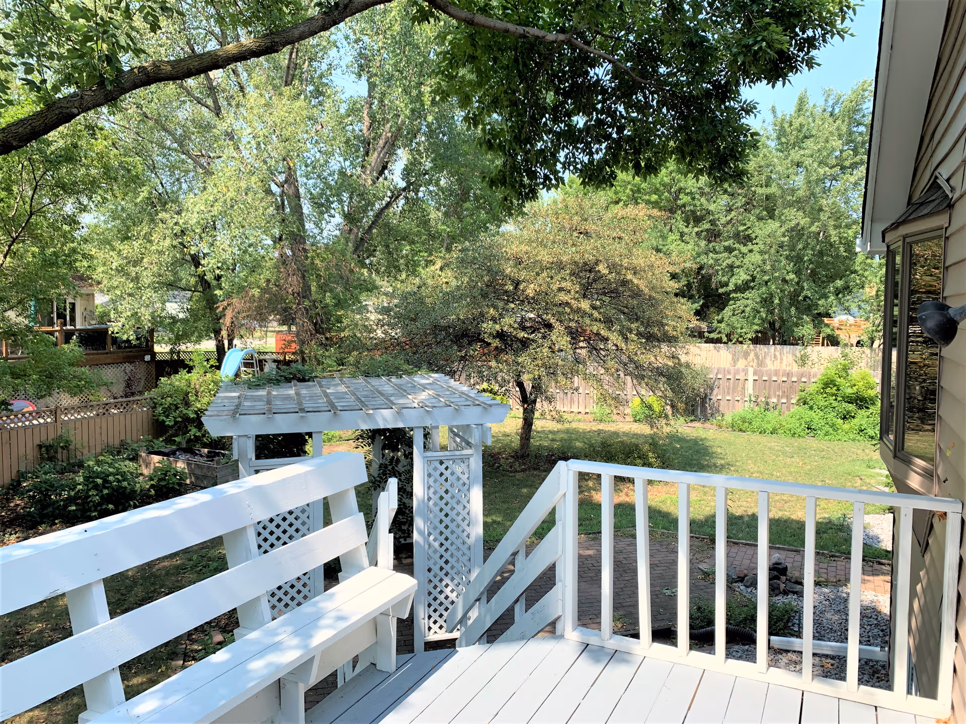 White wooden deck overlooks a backyard with trees, a pergola, and a fenced yard.