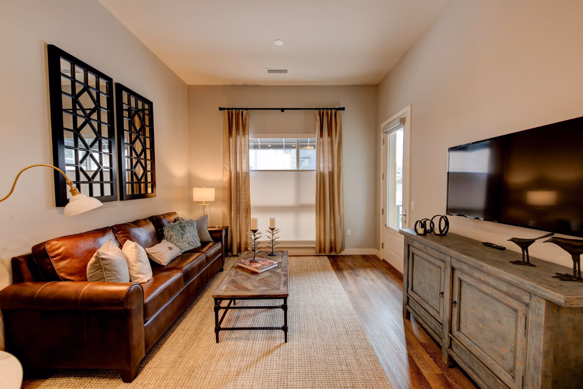 Cozy living room with a brown leather sofa, coffee table, large TV on a wooden console, and a window with curtains.