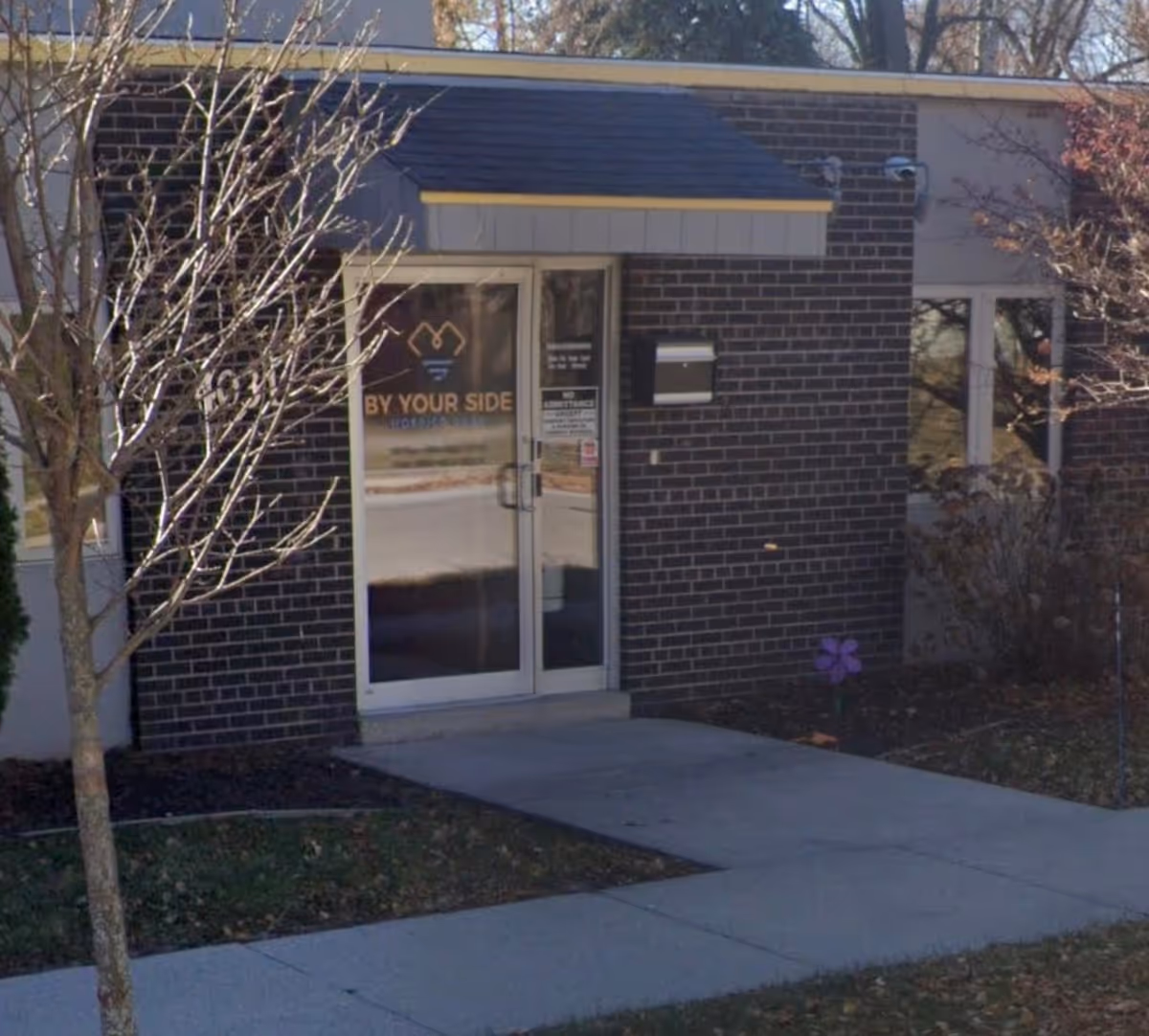 Brick building entrance with glass doors and 'BY YOUR SIDE Hospice Care' signage.