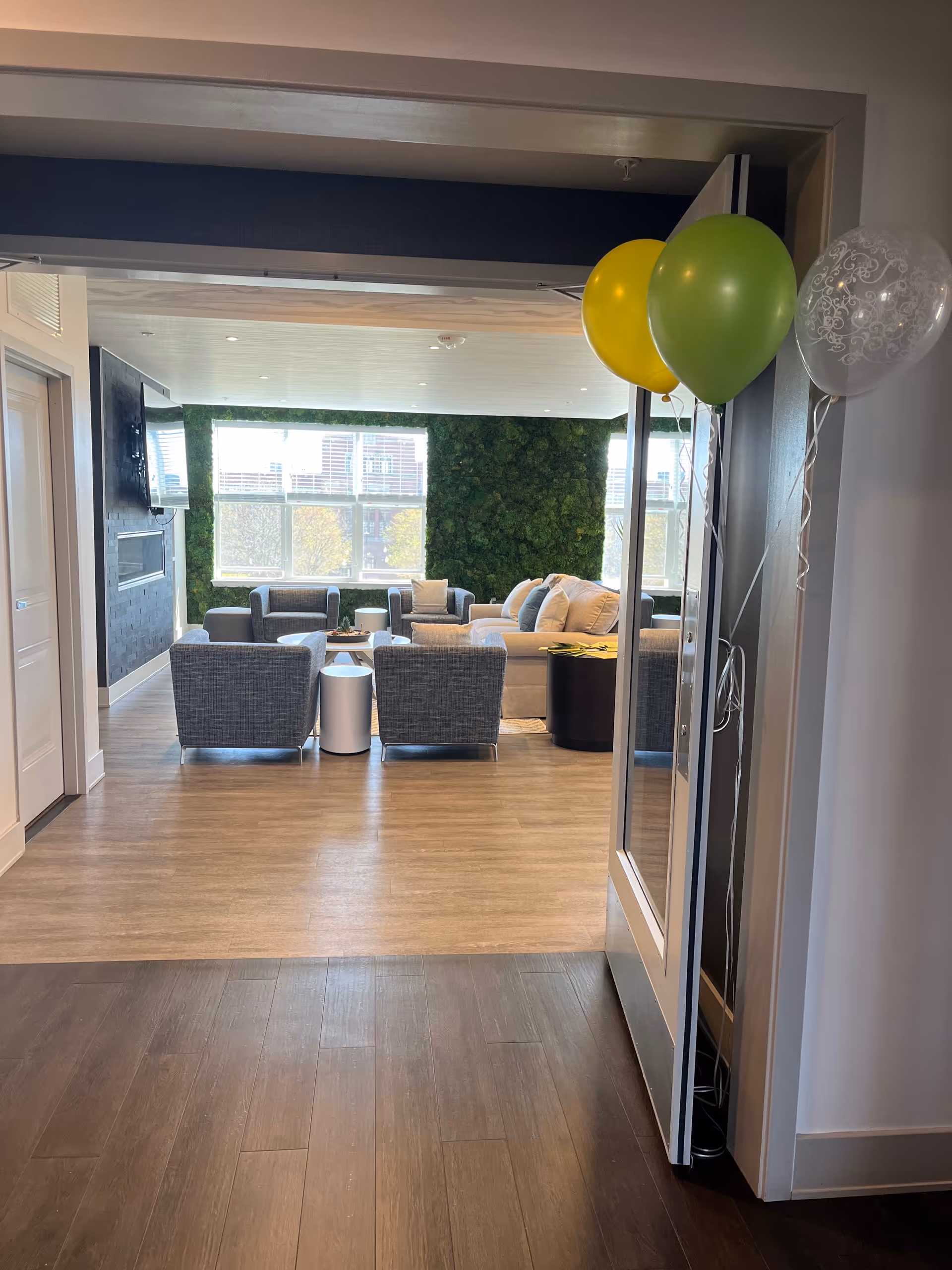Bright senior living lounge with gray seating, coffee tables, green wall, balloons near doorway.