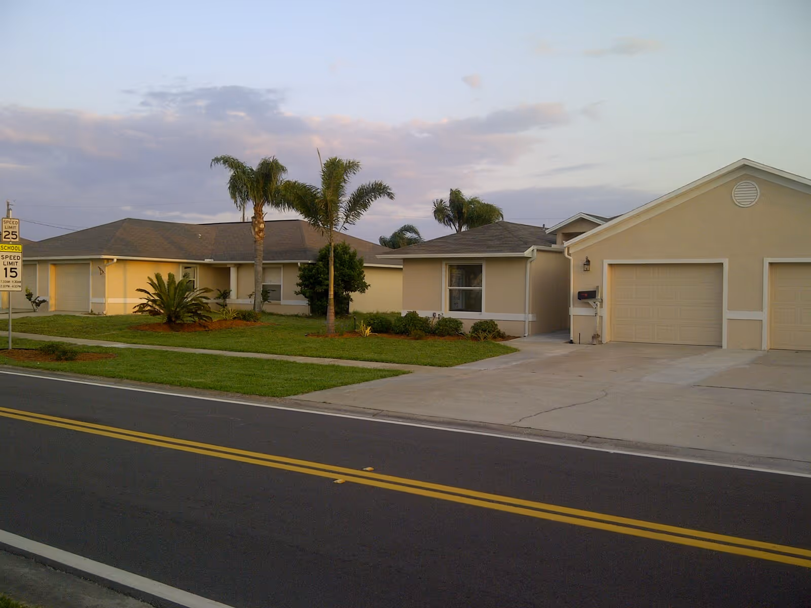 Exterior view of a single-story residential building with beige walls and a gray roof, surrounded by palm trees and green lawn, adjacent to a paved road with yellow dividing lines and a speed limit sign.