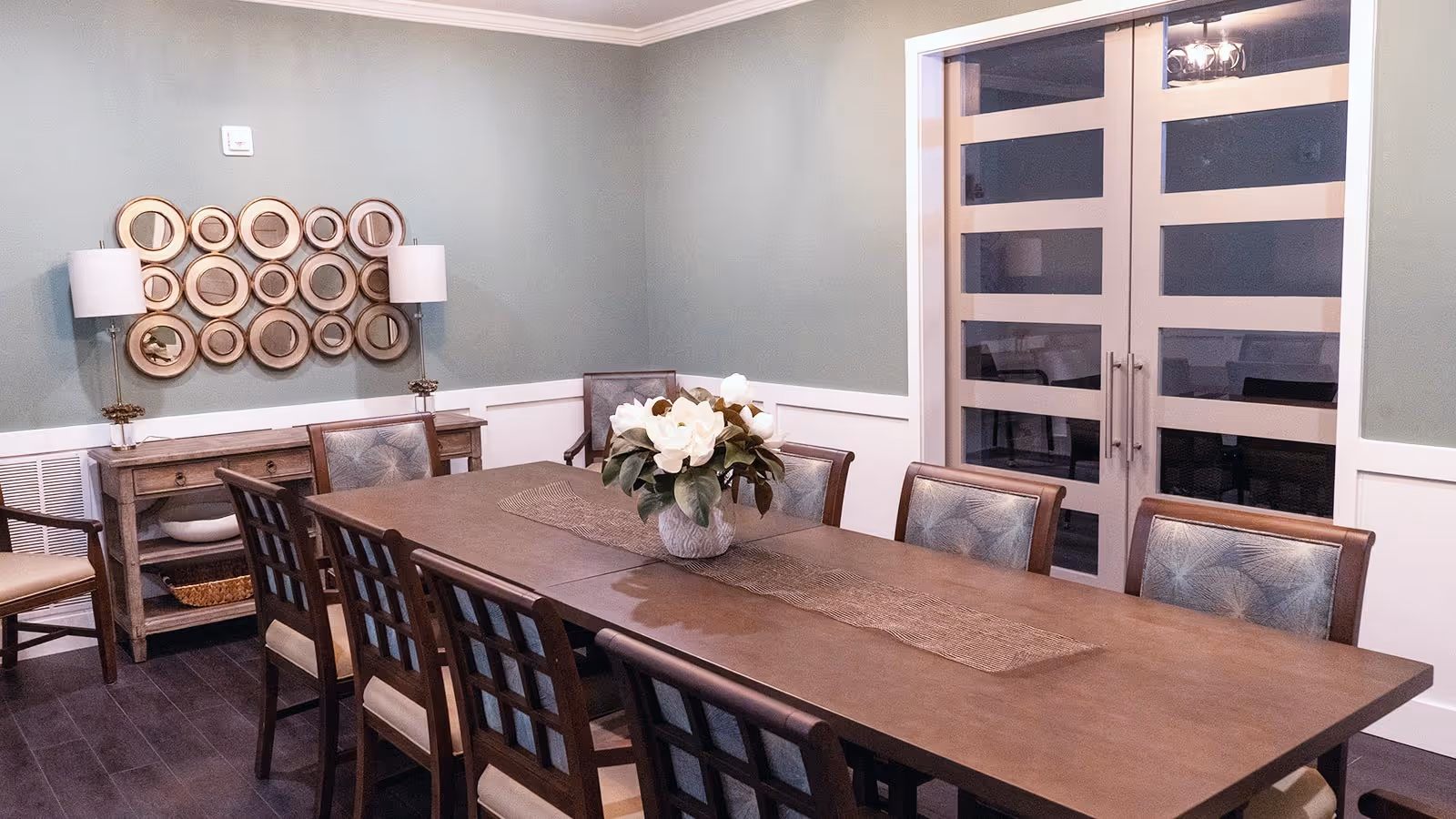 Long wooden dining table with chairs and a floral centerpiece in a softly lit dining room with decorative wall art and glass double doors.