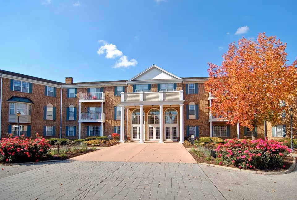 Brick senior living facility with arched entrance, columns, and autumn trees framing the front