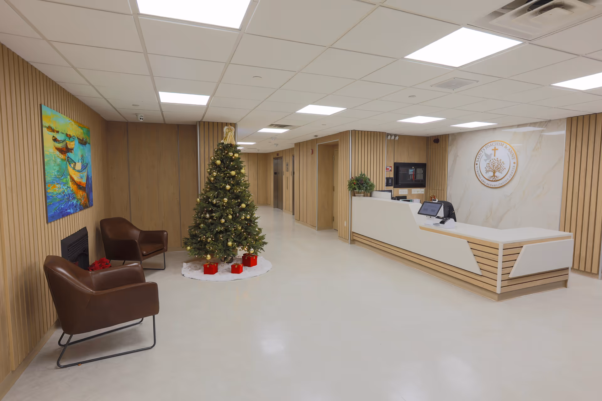 Reception area of Saints Joachim & Anne Nursing & Rehabilitation Center with a decorated Christmas tree in the center, two brown chairs near a wall with a colorful painting, and a modern reception desk with the facility's emblem on the wall behind it.