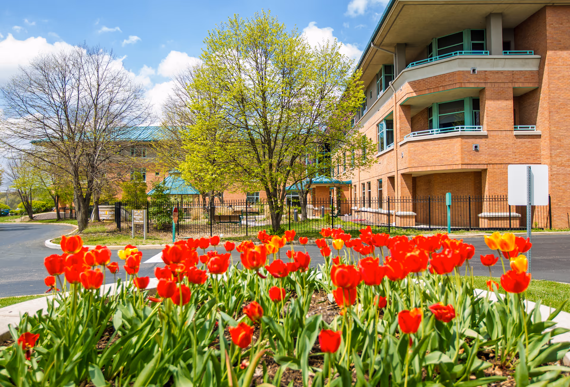 A bright outdoor scene featuring a bed of vibrant red and yellow tulips in the foreground, with a brick building and green trees in the background under a partly cloudy blue sky.