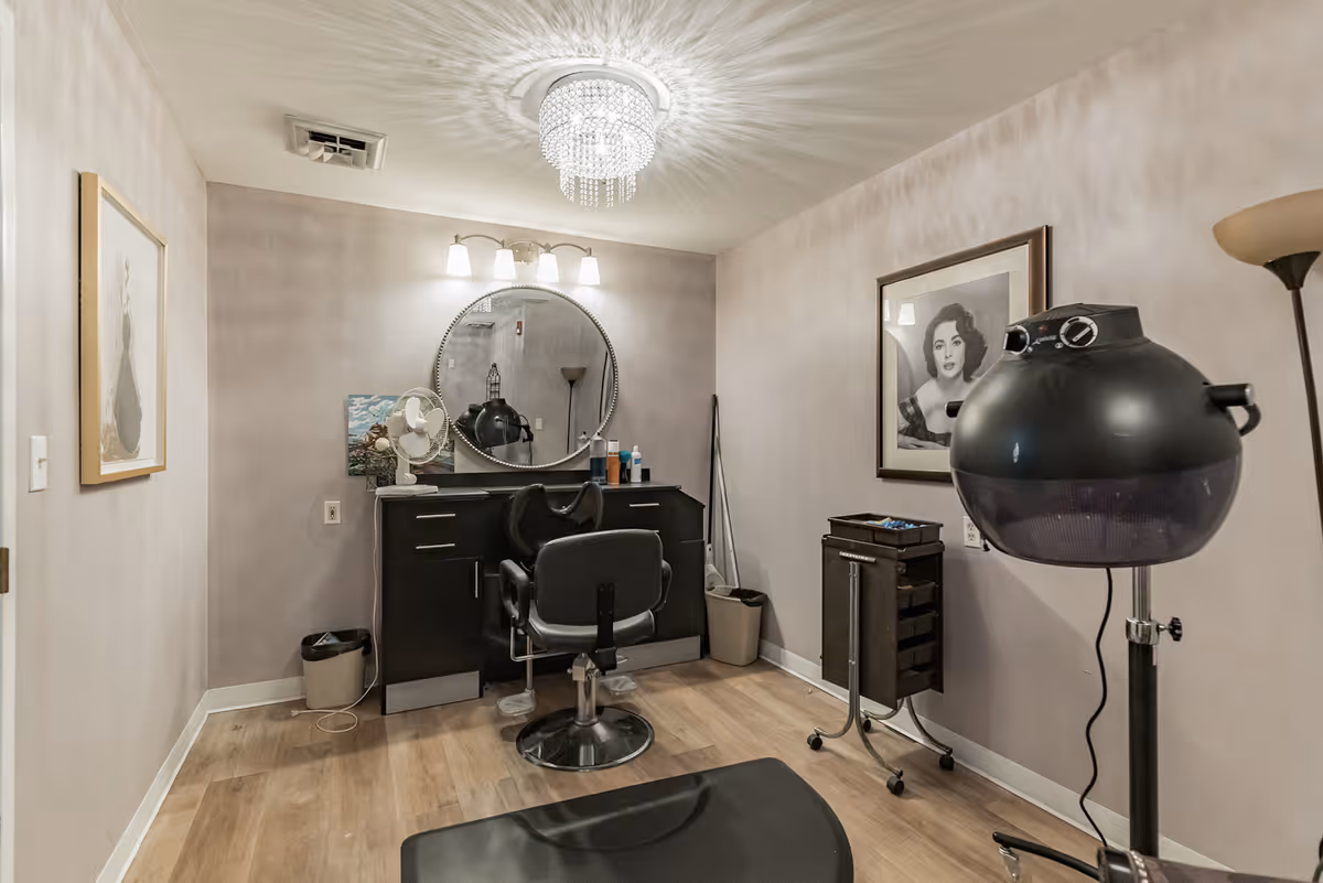 A small salon room with a black styling chair in front of a black cabinet with a round mirror above it. The room has light-colored walls and wood flooring. There is a black hair dryer on a stand, a rolling cart with drawers, a floor lamp, and framed pictures on the walls. A ceiling light fixture with hanging crystals is visible.