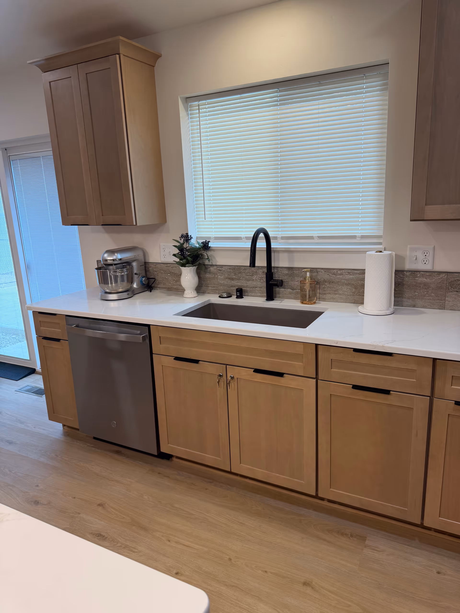 Modern kitchen with light wood cabinets, stainless sink, dishwasher, and window blinds.