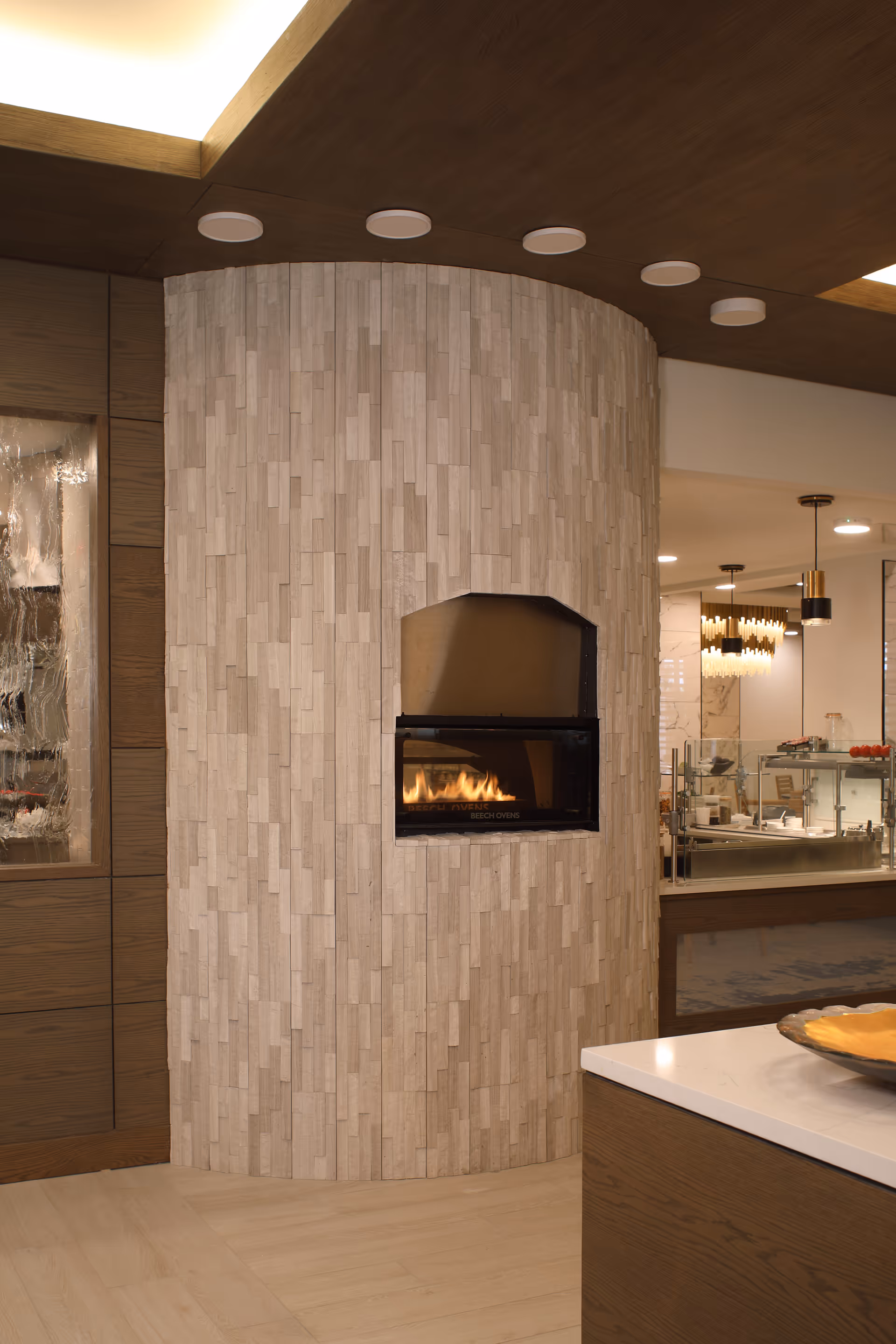 A modern interior space featuring a curved wall with light-colored vertical tile patterns and a built-in fireplace with visible flames. The ceiling has recessed circular lights and wood paneling. To the right, there is a countertop with a decorative bowl and a view into a dining or kitchen area with pendant lights and glass partitions.