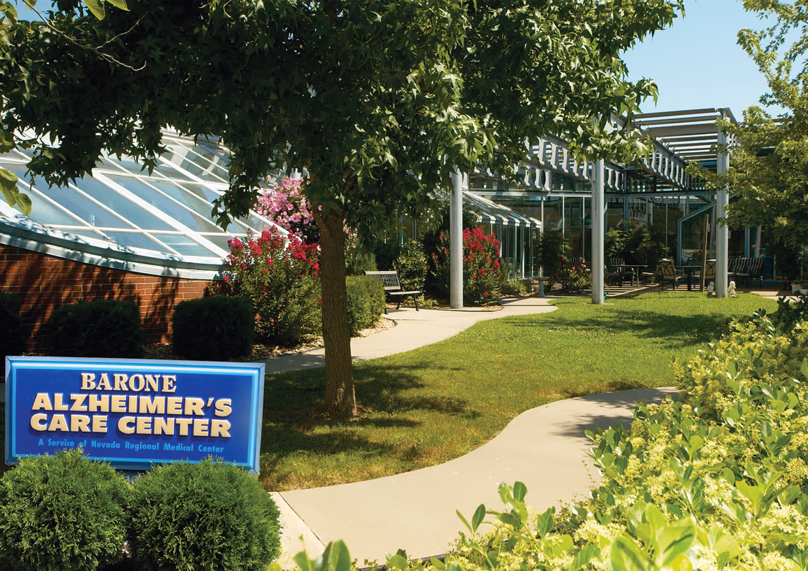 Outdoor garden area at Barone Alzheimer's Care Center with a curved concrete pathway, green grass, bushes, trees, and flowering plants. The building has large glass windows and a pergola structure. A blue sign with yellow and white text reads 'Barone Alzheimer's Care Center, A Service of Nevada Regional Medical Center.'