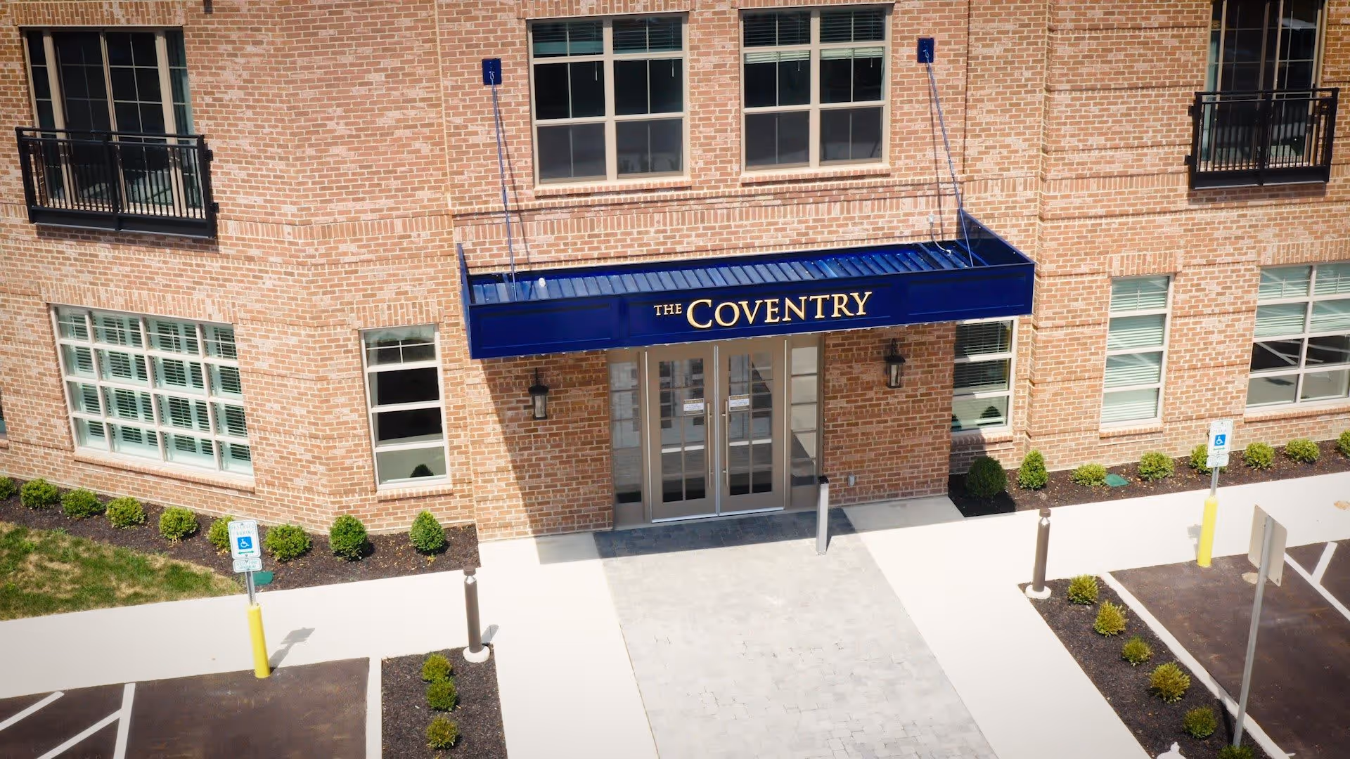 Front entrance of The Coventry building with a blue awning displaying the facility name above double glass doors. The building is made of red brick with several windows and small landscaped bushes along the walkway. There are two handicapped parking spaces visible near the entrance.