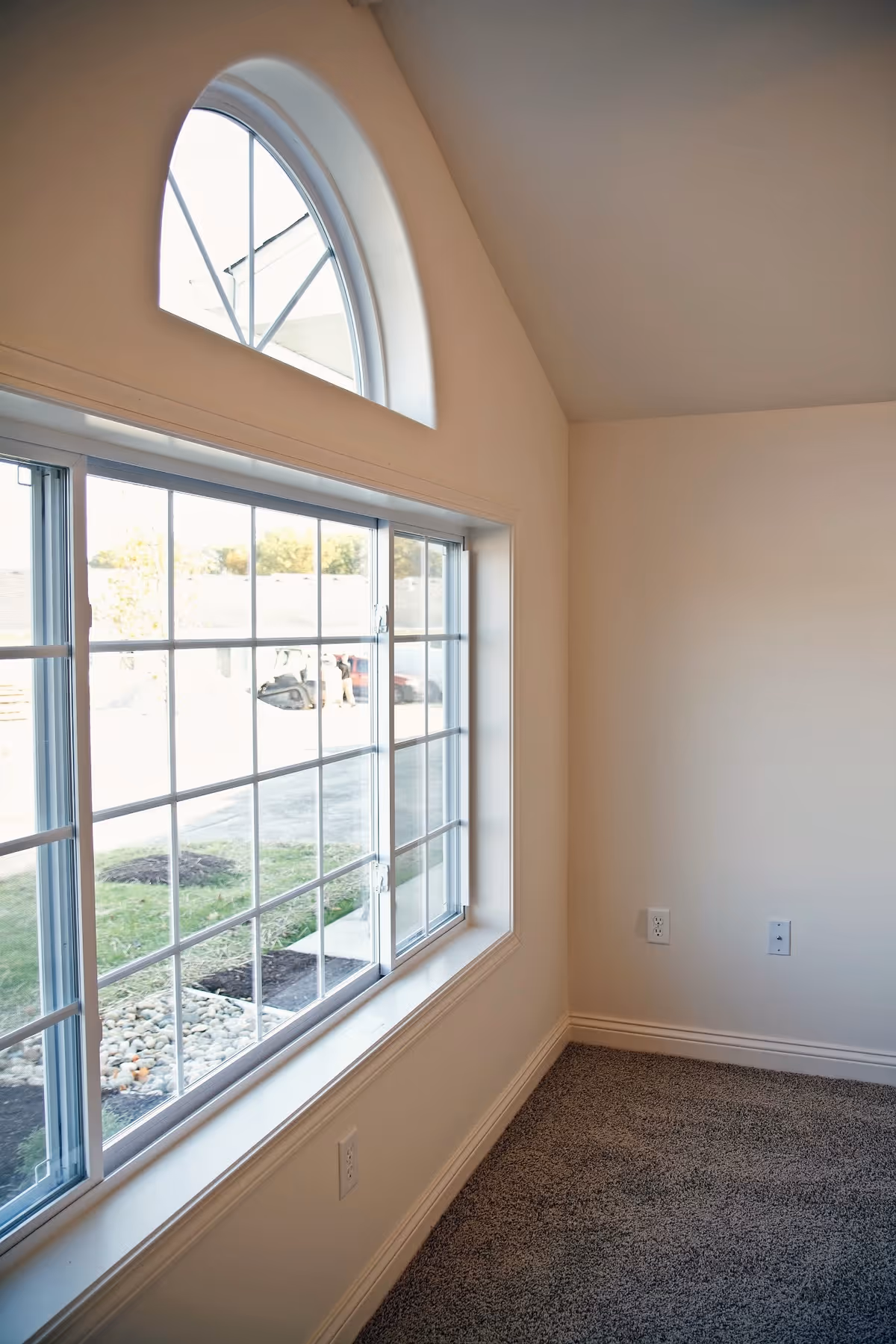 Interior corner of a room with beige walls and carpeted floor, featuring a large rectangular window with grid panes and a smaller arched window above it, allowing natural light to enter.